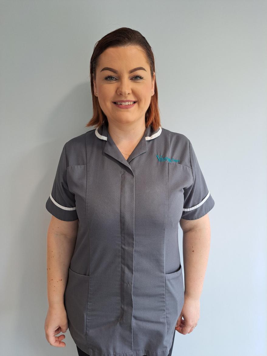 A female carer wearing a grey uniform smiling.