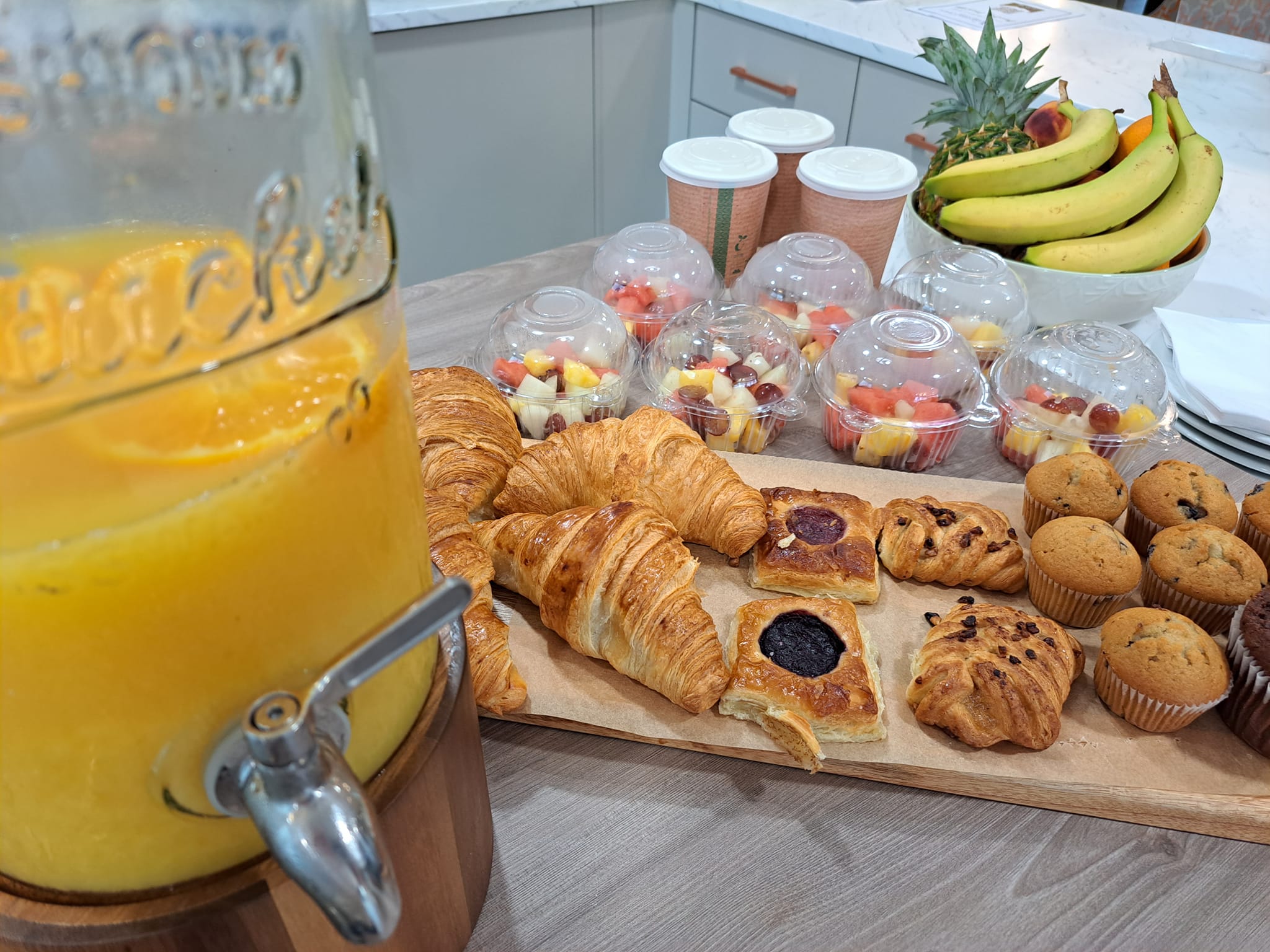 Orange juice in a large container with tap. White fruit bowl. Seven plastic fruit bowls. Three recyclable porridge containers. A selection of sweet treats including, croissant, blueberry muffins, jam tart, pain au chocolat.