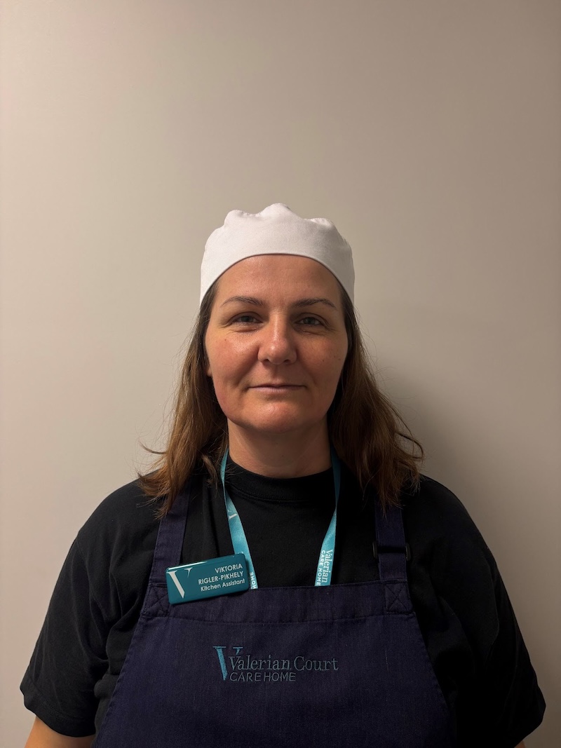 Woman with brown hair and a chefs hat on wearing a black t shirt and navy apron