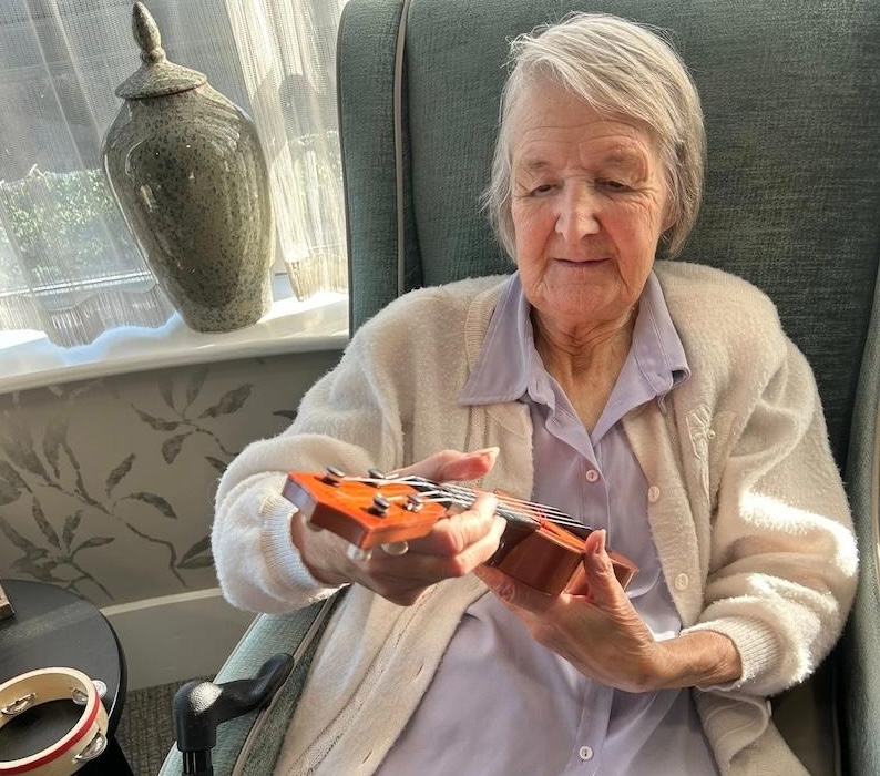 elderly lady sat in an armchair looking at a ukelele in her hand
