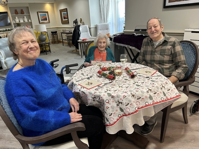 1 elderly man and 2 elderly ladies sat around a dining table at Christmas smiling together